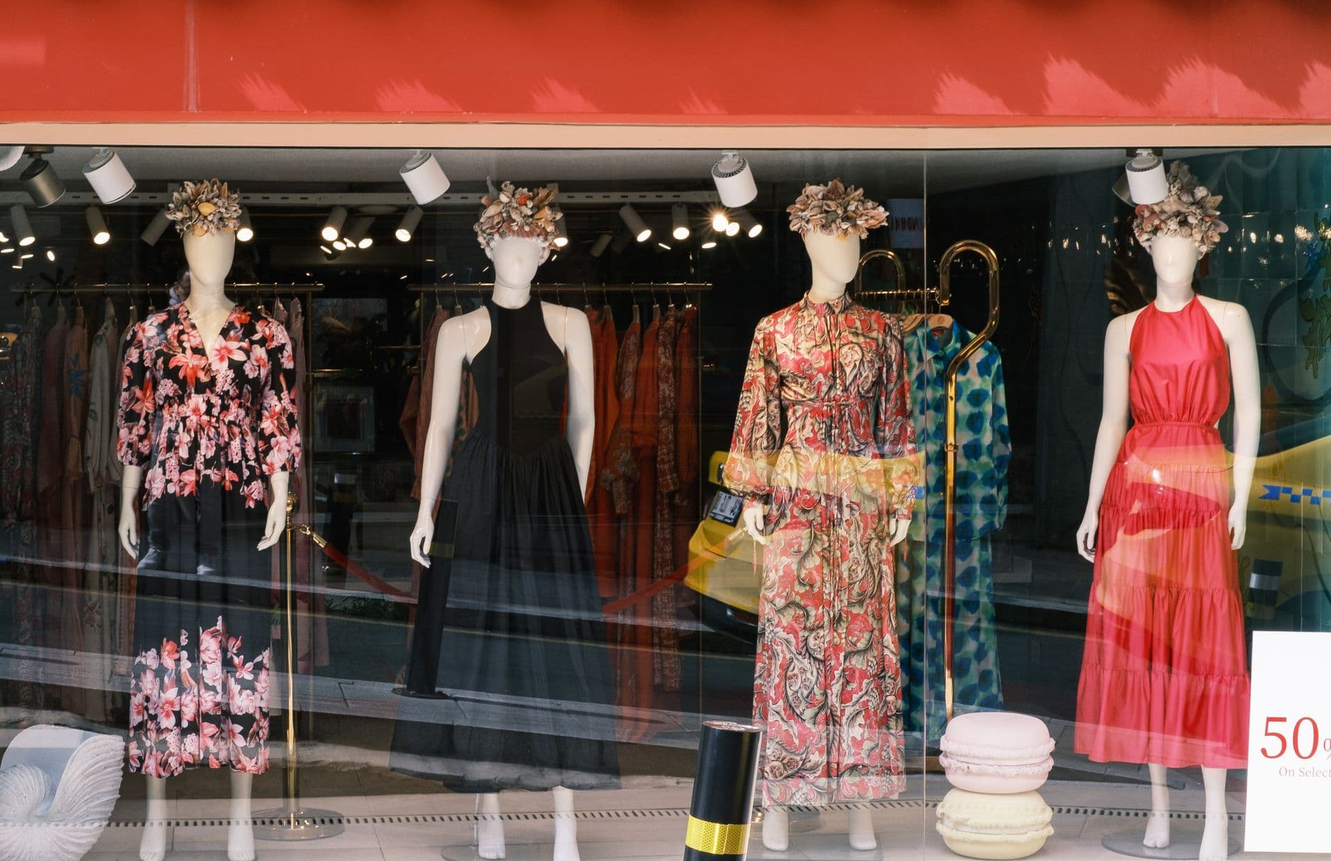 Four mannequins in vibrant dresses and floral headpieces in a stylish window display.
