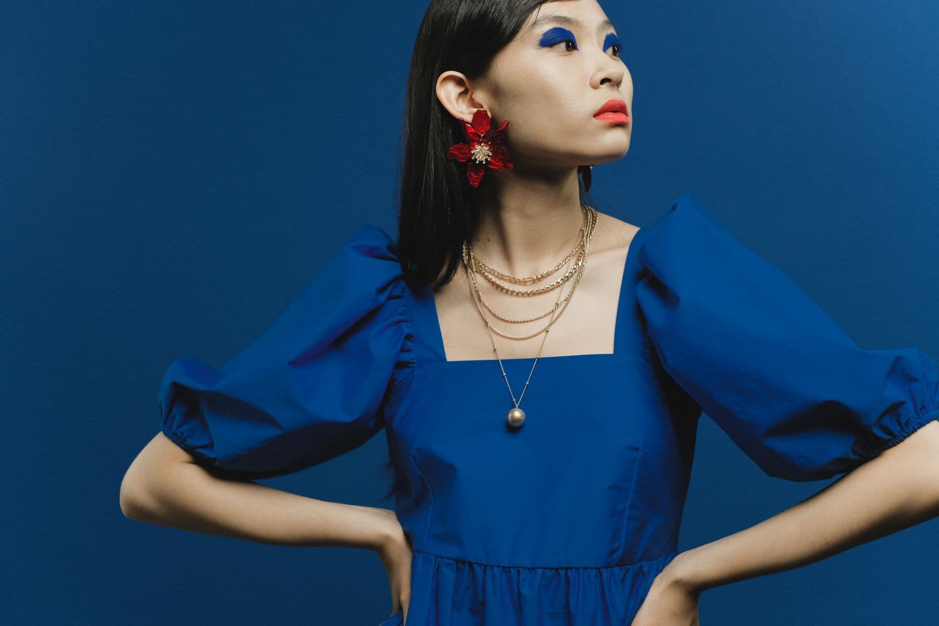 Asian woman striking a pose in a vibrant blue dress with bold eye makeup and red floral earrings.