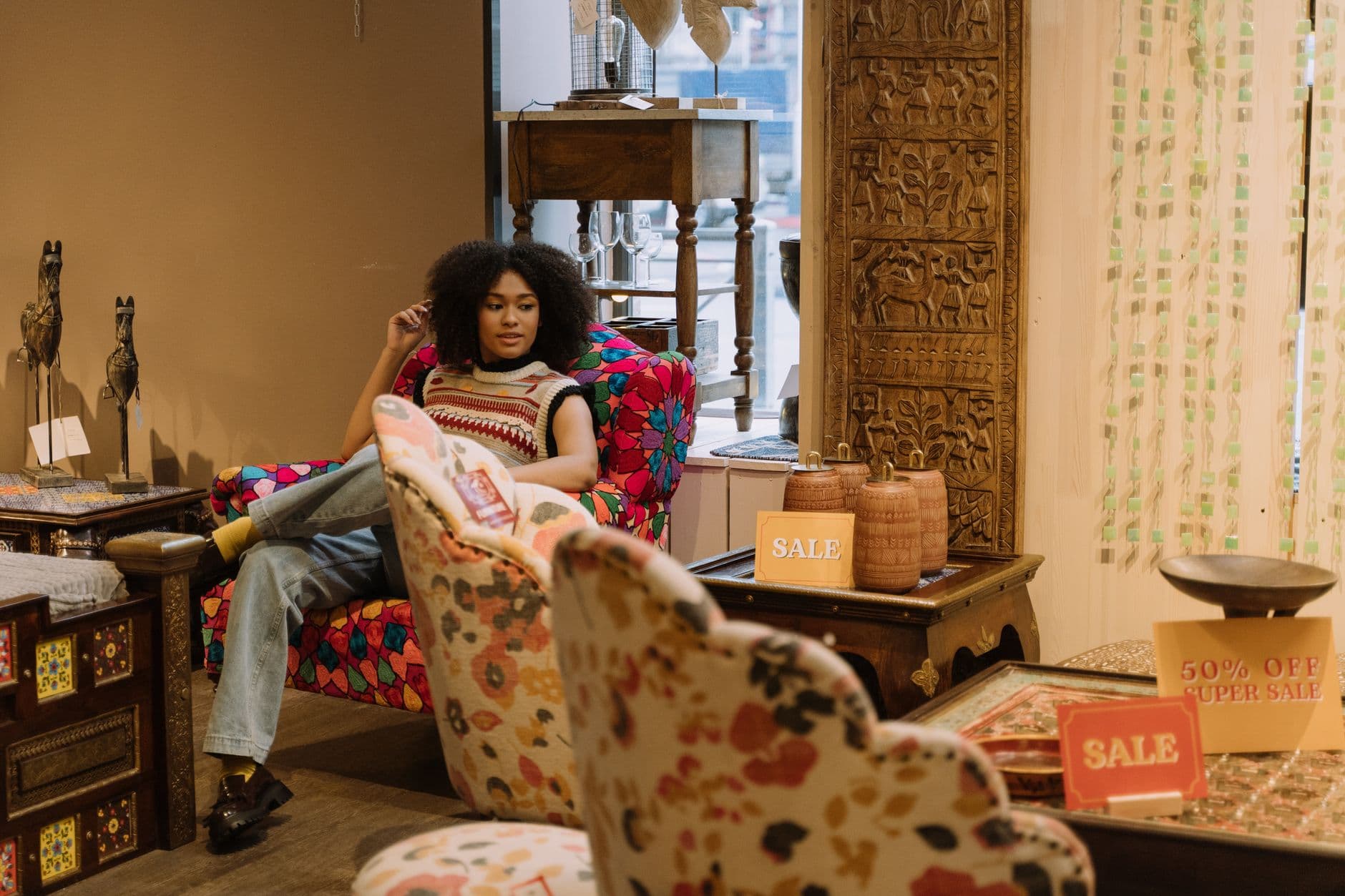 Woman with afro hair relaxing on a colorful chair in a furniture store showcasing sale signs and unique decor pieces.